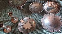 Photo showing several silver plated decorative serving dishes and two candle holders arranged on a green fabric surface. Items have varying designs including pierced, embossed, scalloped edges and pedestal bases. All exhibit tarnishing and need cleaning.