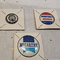 Front side of three vintage 1968 US political pin back buttons for Nixon/Agnew, Humphrey/Muskie and McCarthy campaigns, mounted on white square cards.