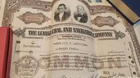 Close up of one stock certificate showing ornate design, portraits, company seal, certificate number, shareholder names, signatures, and date.