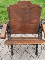 Front view of wooden student chair showing backrest, armrests, and foldable seat with visible wear and scratches on wood, ornate cast iron legs and frame.