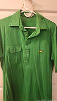 Front view of the green polo shirt showing collar, buttons, chest pocket, and embroidered Augusta National logo.