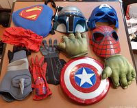 Photo showing assorted superhero masks, gloves, and accessories including Superman logo backpack, Mandalorian, Captain America, Spider-Man masks, Hulk hands, and shield.