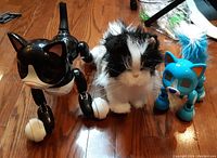 Photo of three electronic cat toys on wooden floor: black and white Zoomer robotic cat, black and white FurReal plush cat, blue Zoomer robotic cat with white tail