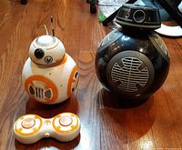 Photo showing BB-8 and BB-9E remote control droids side by side on wood floor with remote controller in front.