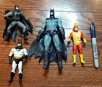 Four DC comic action figures arranged on wooden surface next to Sharpie pen for scale: three Batman figures in different costumes and one Firestorm figure in red and yellow suit with flame head.