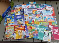 Photo showing a large assortment of colorful children's educational workbooks spread out. Included are titles focused on reading, math, writing, phonics, and language skills for grades pre-K through grade 3.