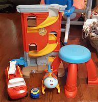 Overall view of red and yellow two-level playset with slides, smiling white helicopter toy, blue and yellow small toy, red boat-like vehicle, and blue round stool with red legs and paw print detail