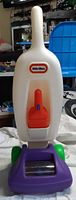 Front of Little Tikes toy vacuum cleaner showing handle, switch, logo, and purple base with green wheels.