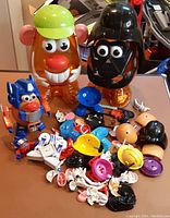 Two large transparent orange Mr. Potato Head storage heads with themed hats and helmets with assorted small parts in front.