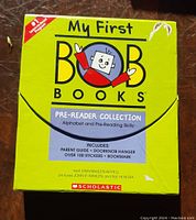 Photo of the yellow box for My First Bob Books Pre-Reader Collection containing reading materials including a parent guide, doorknob hanger, stickers, and bookmark from Scholastic.