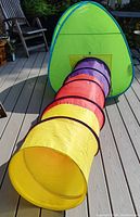 Side view of the green pop-up tent with attached colorful play tunnel extended forward on a wooden deck.