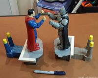 Front view of both Batman and Superman robot fighters on rocky bases showing raised fist boxing posture and colored levers for game operation with a pen for scale.