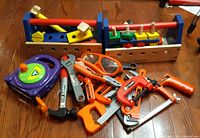 Entire lot arranged on wooden floor showing two Melissa & Doug wooden construction toolboxes along with various orange, grey, black and purple plastic toy tools lying in front.