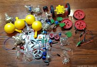 Wide view of assorted vintage Capsela parts scattered on a wooden surface with yellow spheres, clear plastic components, wheels, and wires visible.