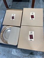 Three unopened boxes containing serving plates with IVV labels and one visible plate with textured gold rim.
