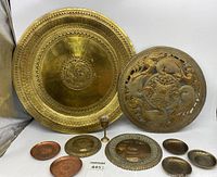 Large brass wall hangings, small decorative plates, coasters and a small goblet arranged on a flat surface for display