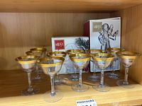 Eleven clear cocktail glasses with wide gold rims displayed on a wooden shelf with Mikasa boxes behind them.