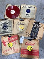 Photo showing several vintage vinyl records with original paper sleeves from Columbia, Capitol, Decca, Victor, Mercury labels. Red vinyl record visible.