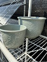 Pair of celadon green planter pots on metal shelf, showing their shape and embossed floral patterns.