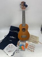 Wooden ukulele with accompanying accessories laid out including bag, picks, instructional materials, and cloth pouch.