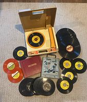 Top view of vintage Fisher-Price children's record player with one 45 RPM record on the turntable, multiple 45s and LPs laid out on floor, two red 45 record holders and red record album.