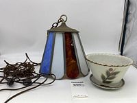 Hanging stained glass light fixture with metal top, blue, white, and amber glass panels, connected chain and power cord, alongside a vintage Otagiri ceramic flower pot with matching saucer.