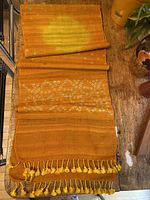 The entire table runner is seen partially folded, showing its tangerine color, cream motifs and fringed tassel ends.