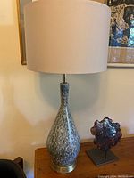 Front view of the tall ceramic lamp on a wooden table, showing the new beige fabric shade and mottled grey-blue and cream ceramic base with brass bottom.
