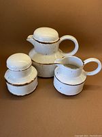 Three pieces including lidded teapot, lidded sugar, and creamer jug arranged against brown background showing cream and cinnamon band design.
