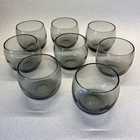 Eight smoky grey roly poly scotch glasses arranged on a white surface, viewed at slight angle showing shape and bulk.