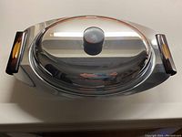 Top-down view of the oval stainless steel serving dish with bakelite handles and lid knob, showing reflective surface and handle colors.
