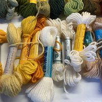 Multiple skeins of wool yarn in yellow, white, and orange colors with labels showing brand names and fiber content.