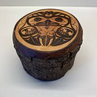 Front angled view showing the wooden round container with bark sides and wood burned hawk faces design on the lid.