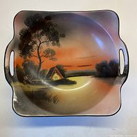 Top-down photo showing the hand-painted scene of a cottage by the lake under a sunset sky with cut-out handles on the bowl sides.