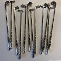 Overview of all 12 vintage silver plated golf club stir sticks showing different types including drivers, irons, putters, and wedges with detailed heads and grip patterns.