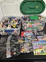 View of two clear plastic containers filled with assorted Lego bricks and parts, a large green Lego baseplate on top of one tub, and several instruction manuals spread in front.