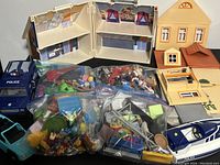 Photo showing the two foldable Playmobil houses, the police vehicle, the blue convertible car, and multiple bags containing Playmobil figures and accessories grouped together.
