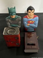 Front view of Batman and Superman ceramic coin banks and two metal bank designs on table.