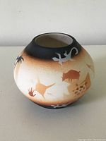Photo showing earth-tone pottery vase with animal and symbolic Native American designs including a lizard and a bison on one side, rounded shape with black top rim.