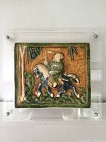 Front view of the antique Chinese ceramic plaque showing a rider on a horse framed with greenery in an acrylic case