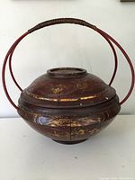 Full frontal view of the antique Chinese wooden lacquer rice basket showcasing round shape, red lacquer surface with cracked areas and gilt painted floral decoration. The circular handle is wrapped in dark material.