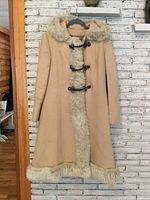 Front view of vintage full length wool coat with fur trim on hood, front, and hem. Shows toggle leather straps and buttons. Coat is beige with visible wear.