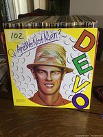 Front cover of the Devo album 'Are We Not Men? We Are Devo!' showing yellow background, stylized man's face with bucket hat, colorful DEVO letters vertically on the right, and handwritten album title on the top left.