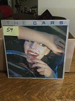 Front cover of The Cars LP album showing a smiling woman behind a car windshield with a yellow sticker marked 59.