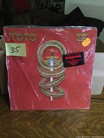 Front album cover of Toto IV showing large sword logo and stickers indicating price and song Rosanna.