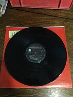 Full view of the black vinyl record atop a red paper sleeve, showing the record surface and label side 1.