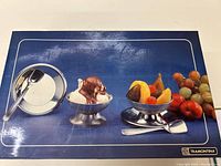 Box lid image showing 2 stainless steel ice cream bowls, a round plate with a spoon, and fresh fruit decor on a blue background. Brand Tramontina visible on bottom right corner.