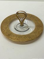 Top-down view of the round clear glass serving tray showing the central handle and wide gold embossed rim with floral pattern.