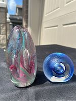 Two art glass paperweights on a black surface outdoors with clear sky background; one tall oval shape with multicolor internal design, one smaller blue circular one.