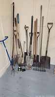Overall view of various garden tools including shovels, rakes, hoes, and hand cultivators arranged in corner against wall on wood floor.